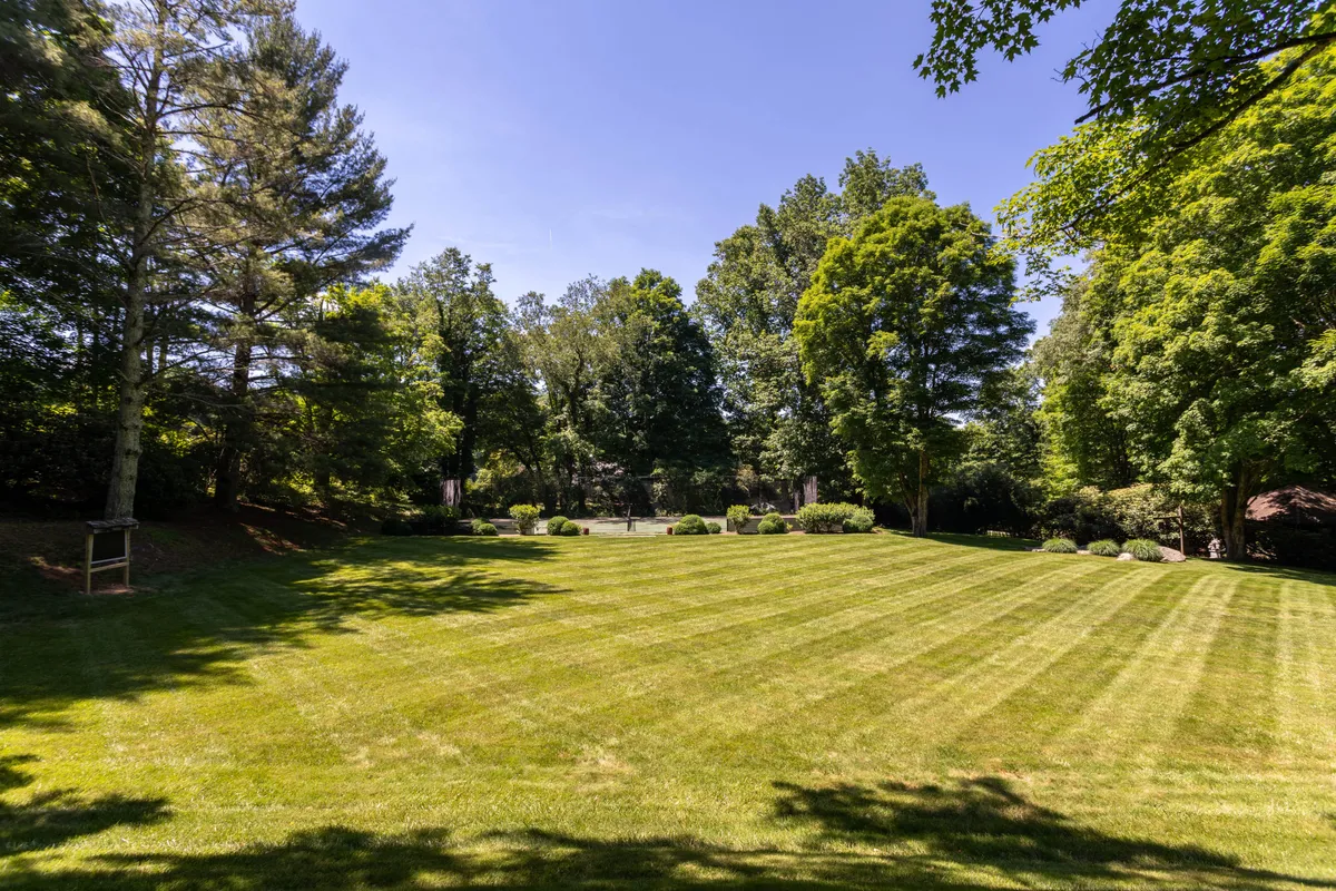 A broad, manicured lawn bordered by tall trees and a lush garden area. The wide-open grassy space is perfect for outdoor games and group activities, evoking the classic summer camp experience. The tranquil setting is bathed in natural light, inviting families and groups to play, picnic, or simply enjoy the outdoors.
