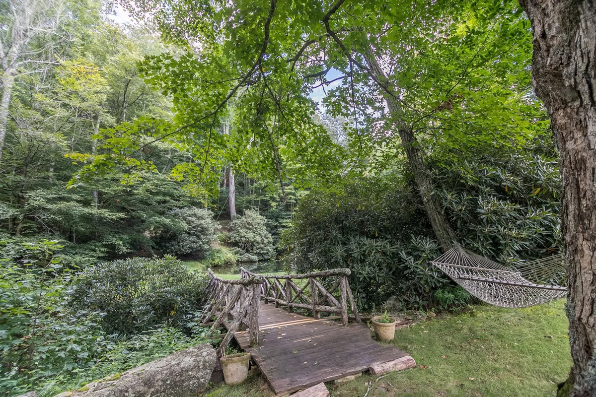 A lush, green outdoor scene featuring a hammock strung between trees beside a charming wooden bridge. The area is surrounded by greenery, providing a serene space for unwinding after a day of activities. The hammock and bridge evoke relaxation and connection with nature.