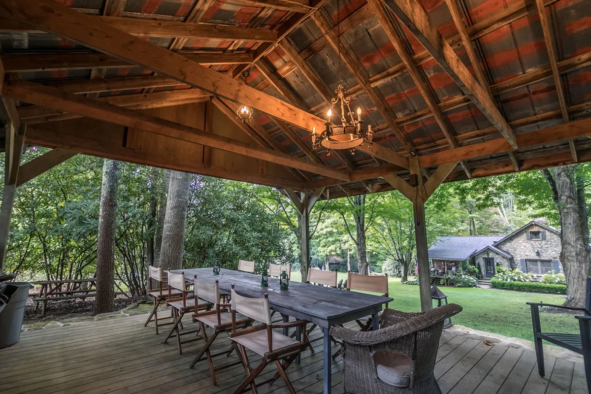 A cozy, covered outdoor pavilion with a large wooden dining table set beneath a chandelier and rustic beams. Surrounded by greenery, this space is perfect for group dining or relaxing by the fireplace after a day's play.