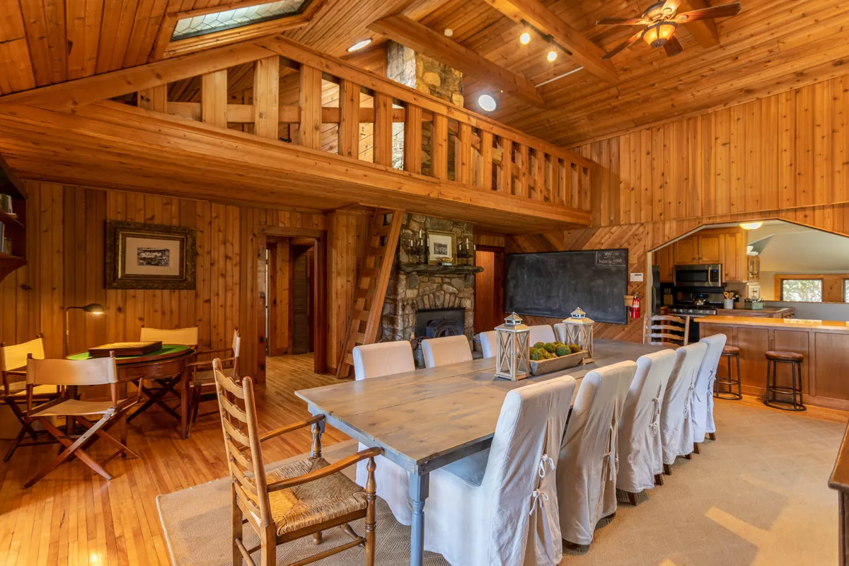 This image features a spacious, communal dining area with a large table capable of seating many, a stone fireplace, open kitchen views, and exposed wooden beams. The table is set for togetherness, while the kitchen in the backdrop ties in culinary preparation with shared dining experiences.