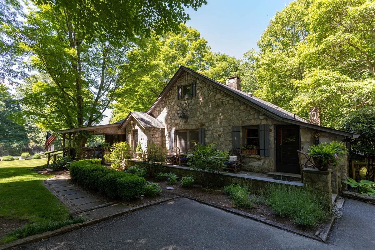 This image features the stone facade of the house, surrounded by lush greenery, with a front porch that has inviting seating and an American flag displayed. The scene is bright and welcoming, emphasizing the connection between the home and its natural mountain surroundings.