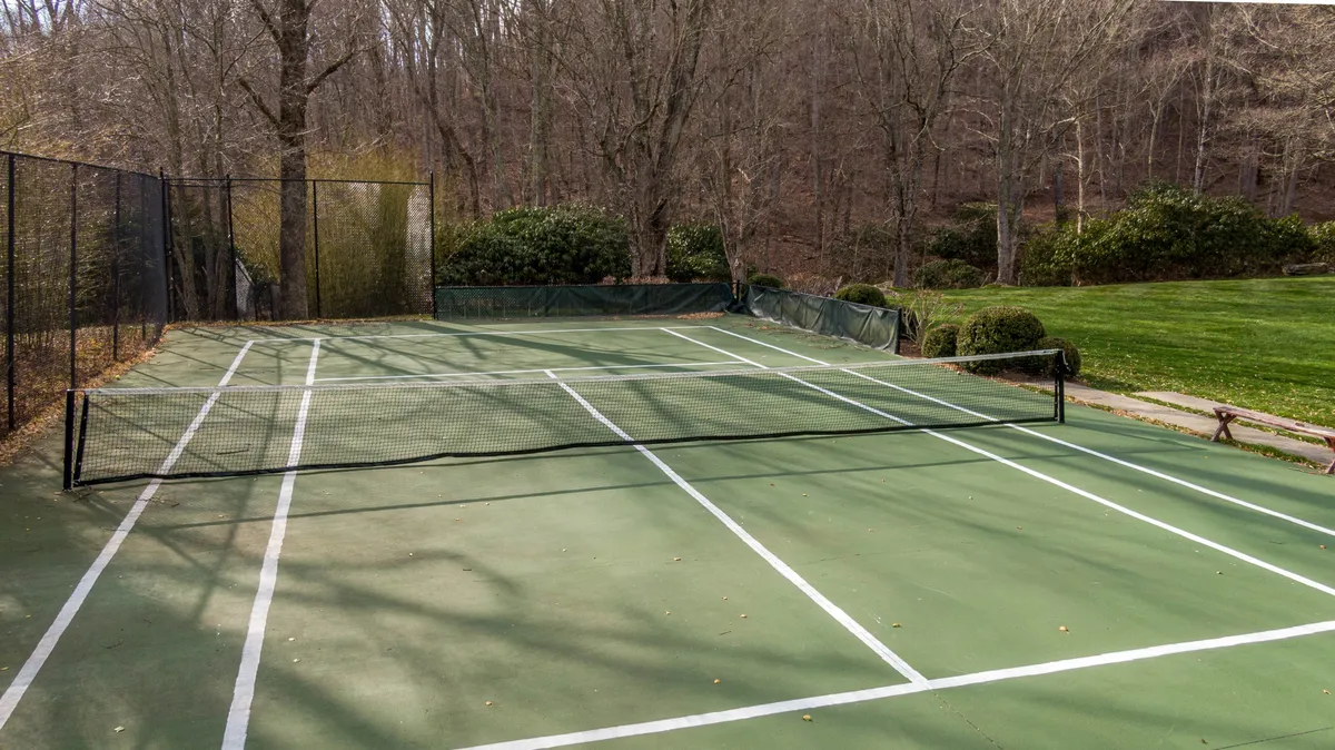 A fenced tennis court surrounded by leafy trees and lush greenery. The court is bright, inviting, and clearly set up for friendly matches or group tournaments, with adjoining grassy areas suitable for spectators or picnics.