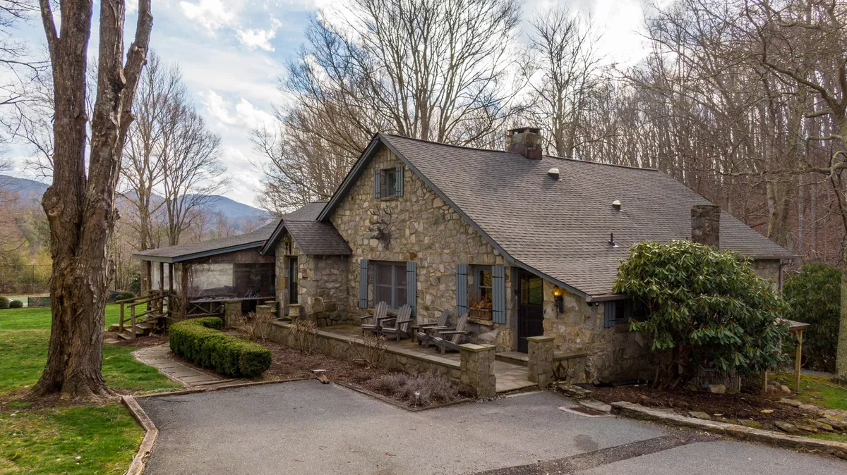 This image shows the stone facade of Beech Haven with ample front porch seating, surrounded by lush trees and with a scenic mountain backdrop. The inviting porch and expansive green space capture the property's connection to the mountain setting and its year-round outdoor appeal.