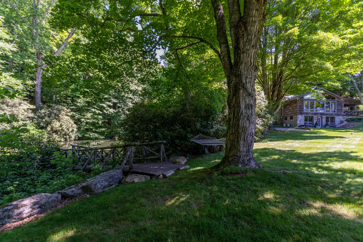A tranquil exterior featuring a large tree, a wooden bridge, the house with expansive windows, and lush greenery. The scene evokes the peacefulness of nature and opportunities for outdoor picnics, morning coffee, and walks around the property throughout all seasons.
