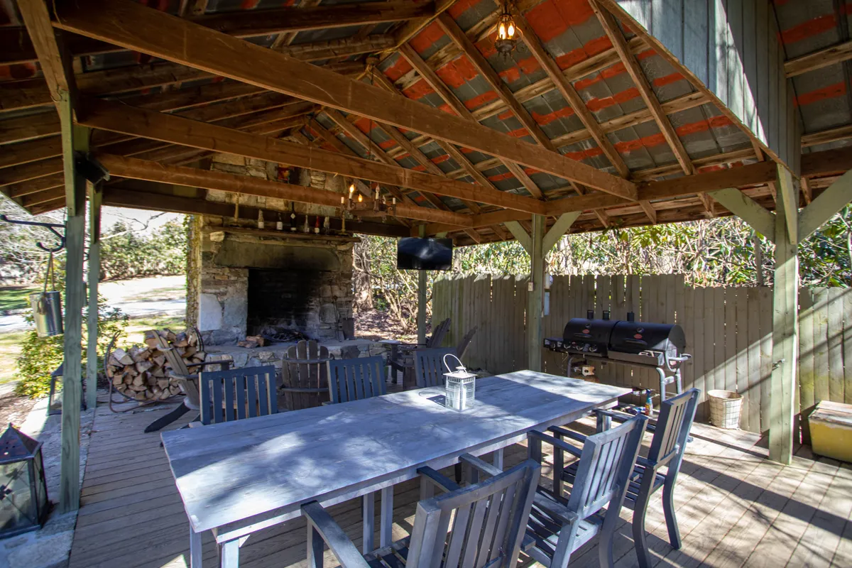 A spacious covered outdoor pavilion with a large dining table, grill, and fireplace, all illuminated by soft overhead lighting. The area is designed for group gatherings, outdoor meals, and fireside storytelling, with a relaxed and welcoming ambiance.