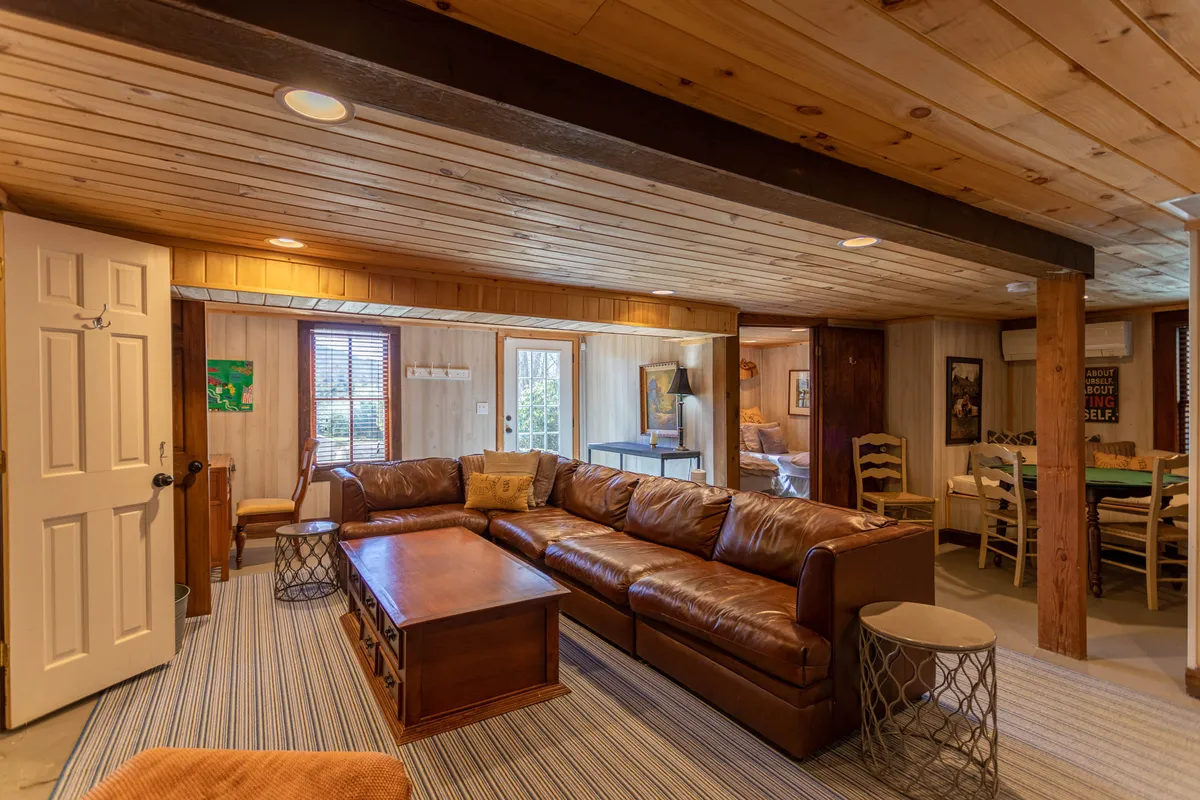 A welcoming lower-level lounge with a large leather sectional sofa, a coffee table for board or card games, wooden beams, and a dining table visible in the background. The space radiates warmth, ideal for movie nights or poker tournaments.