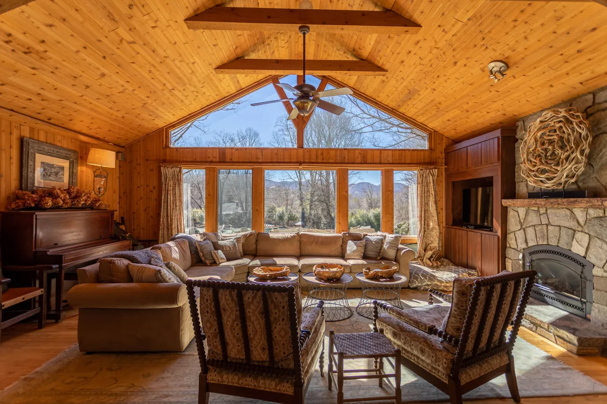 A spacious living room with a large sectional sofa facing a stone fireplace, framed by panoramic windows that flood the room with natural light. Exposed wooden ceiling beams and a piano add warmth and charm, while plush seating invites relaxation and group gatherings.