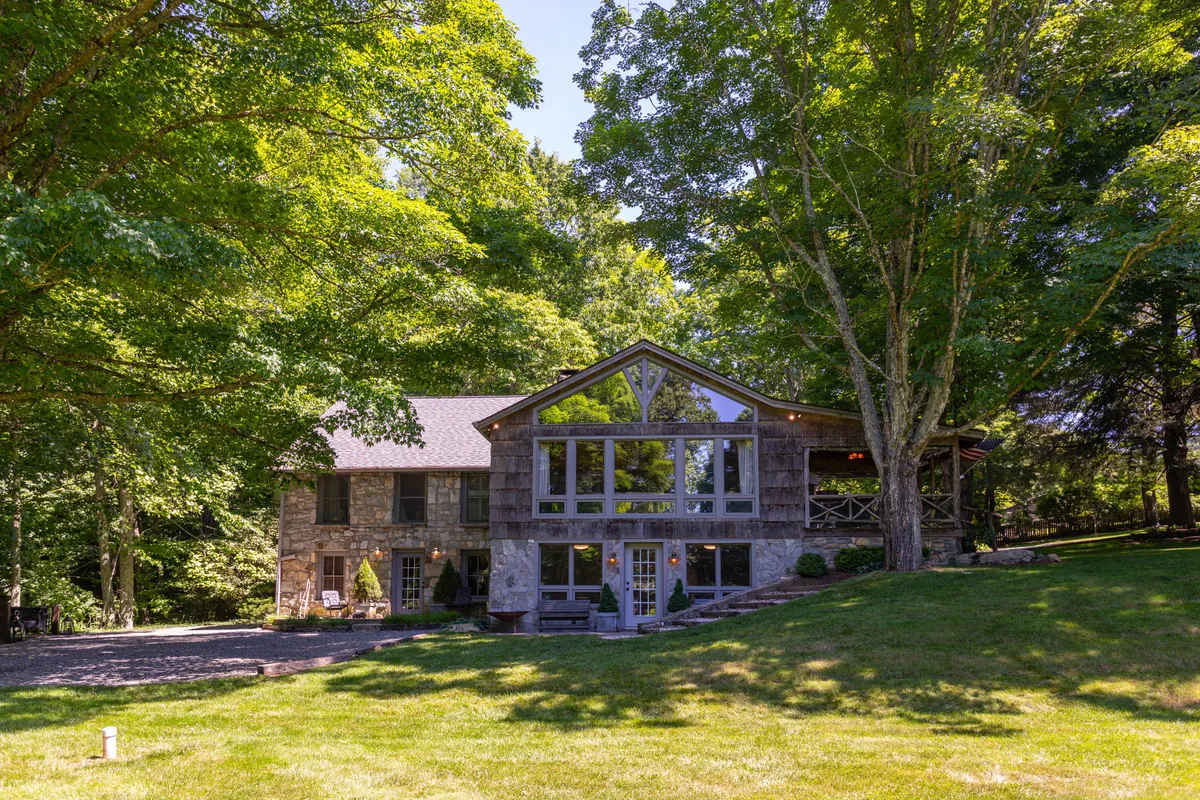 This exterior shot focuses on the house's large windows, stone facade, surrounding trees, and an open lawn area. The ample yard space invites team sports, outdoor gatherings, and playful activities, while the inviting porch and green surroundings emphasize the property's connection to mountain living.