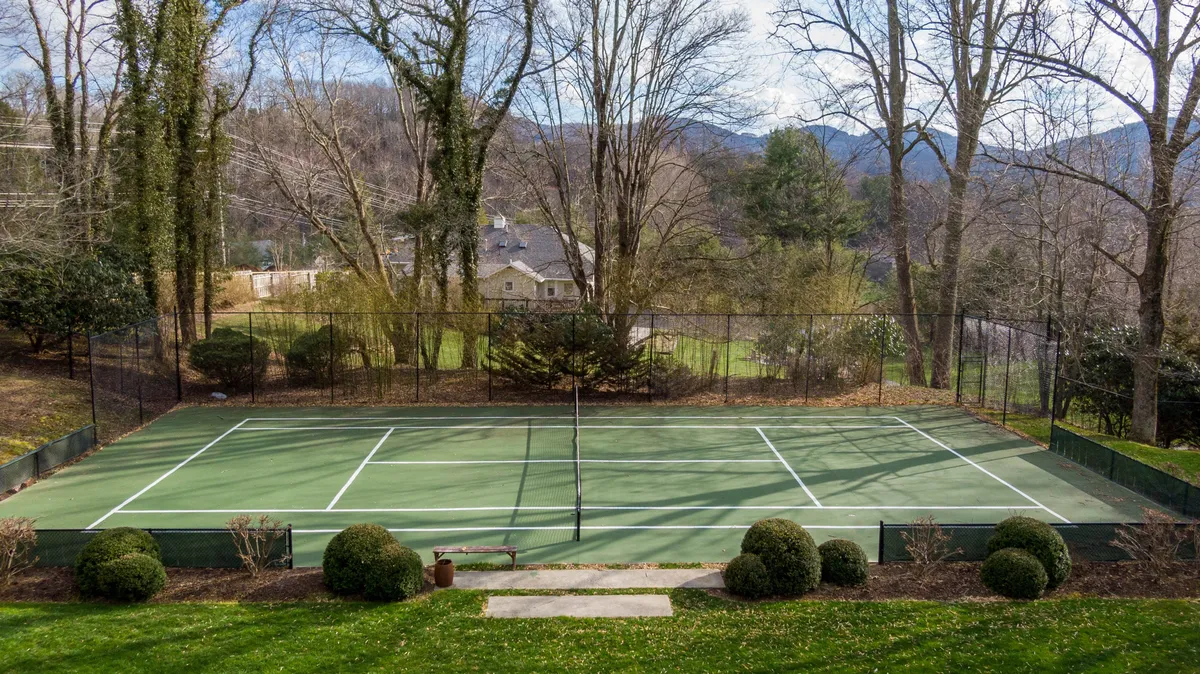 An inviting exterior shot of the private tennis court surrounded by lush trees and set against a mountain backdrop. Benches on the sidelines invite spectators or a moment of rest after a match.