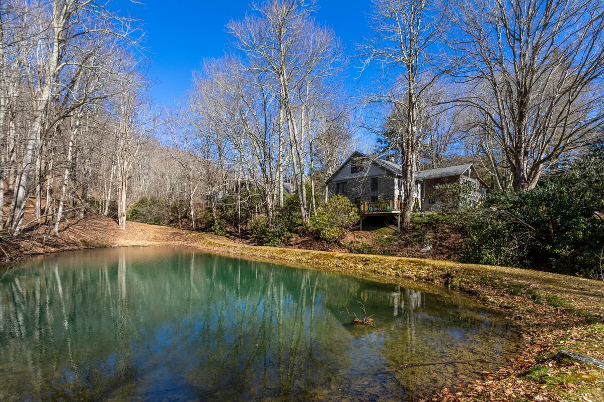 This image captures a tranquil, sunlit pond surrounded by towering trees with the house visible in the backdrop. The serene water invites fishing, while the spacious setting hints at opportunities for nature walks or outdoor relaxation. The golden hour lighting adds to the inviting outdoor atmosphere.