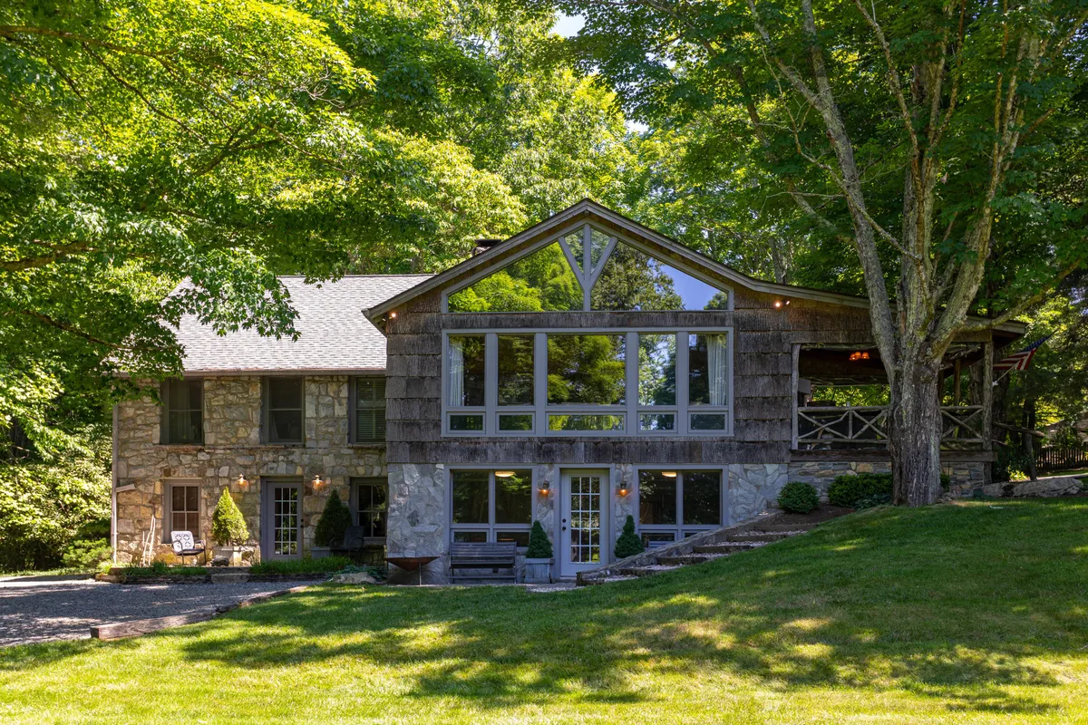 The image showcases the exterior of a beautifully restored stone and wood single-family house surrounded by lush greenery. Large glass windows reflect the golden hues of sunset, and a spacious porch invites relaxation. The expansive yard hints at space for gatherings and play, nestled within a serene mountain setting.