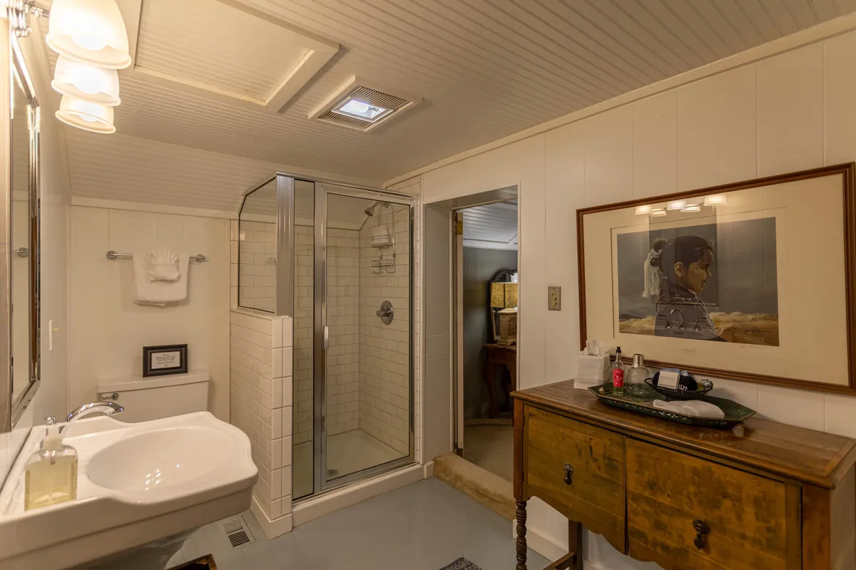 A modern bathroom with a glass shower enclosure, wooden dresser, framed artwork, and a bright sink area. The space feels serene, organized, and designed for comfort, suggesting both privacy and luxury.