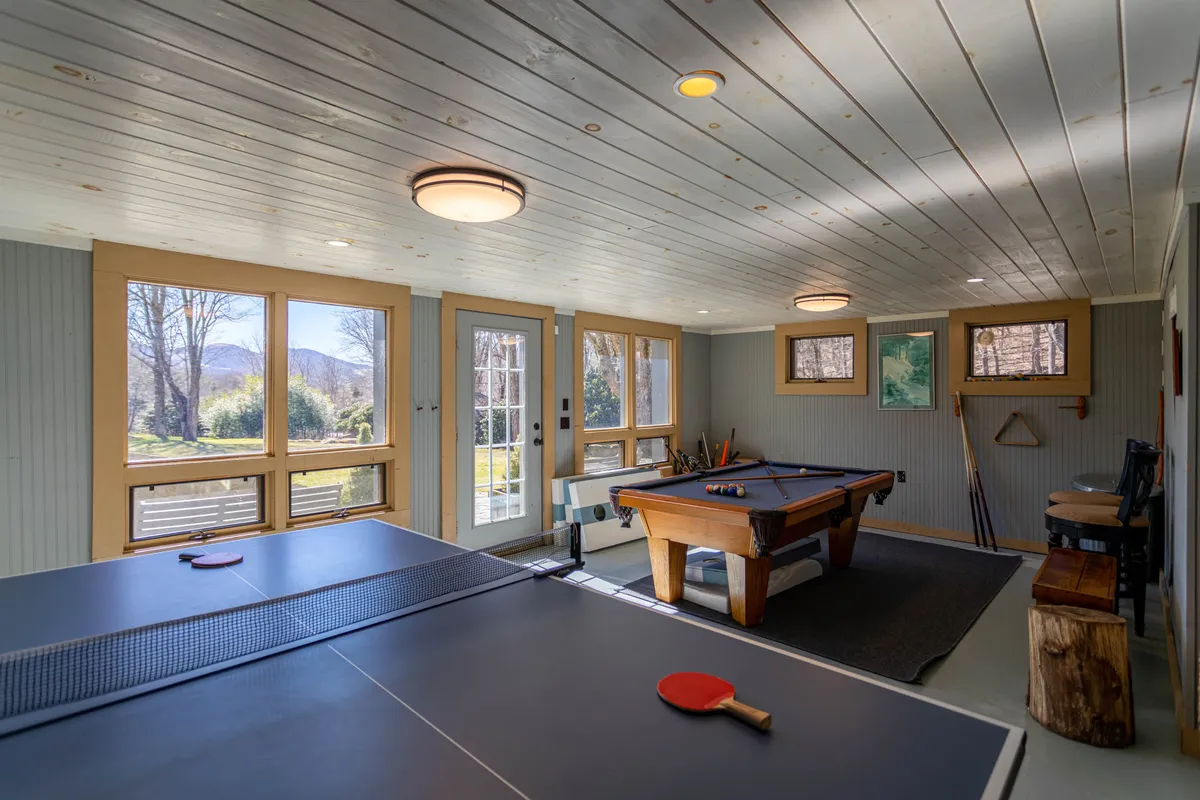 A bright and spacious game room featuring a pool table and ping pong table, complemented by large windows that let in natural light and offer views of the outside. Comfortable seating invites guests to relax between games.